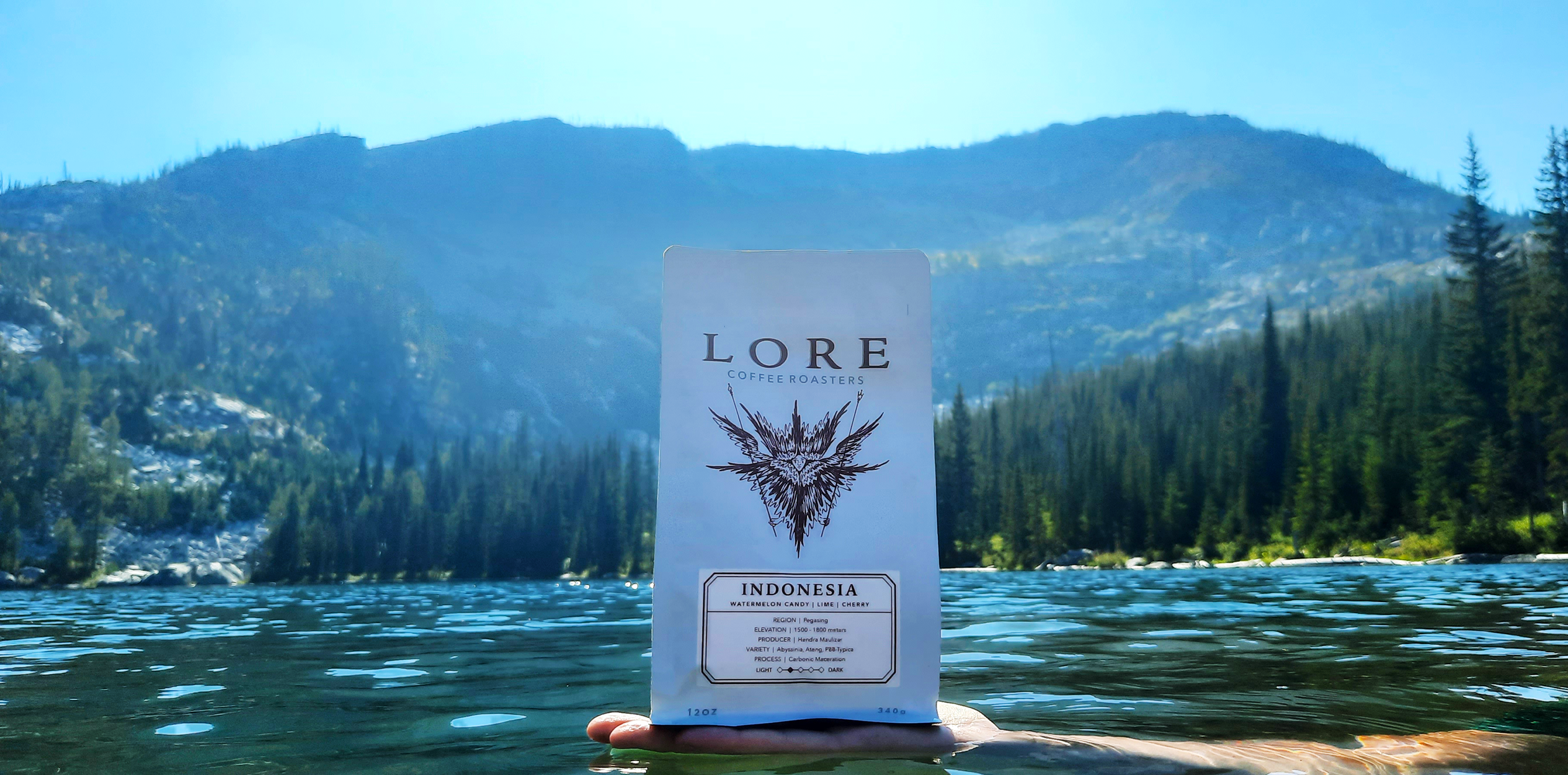 A bag of Lore's Indonesia Coffee being held on a person's hand in a lake with a backdrop of mountainous terrain covered in pine trees.